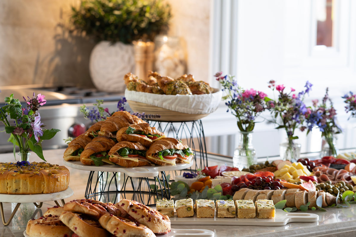A buffet-style brunch table food spread on a table with croissants, sandwiches, bagels, sliced bread, fruit, cheese, and small flowers in vases in a bright indoor setting.