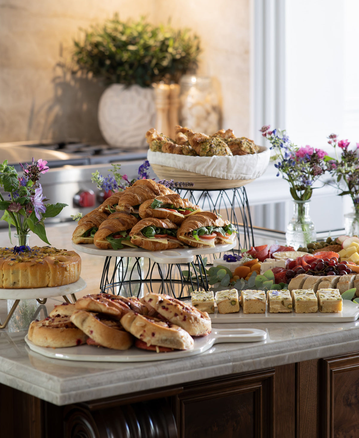 A brunch catering table of assorted pastries, croissants, sandwiches, fruit, and small cakes arranged on tiered stands and platters on a kitchen counter, with flowers and a softly lit interior in the background.