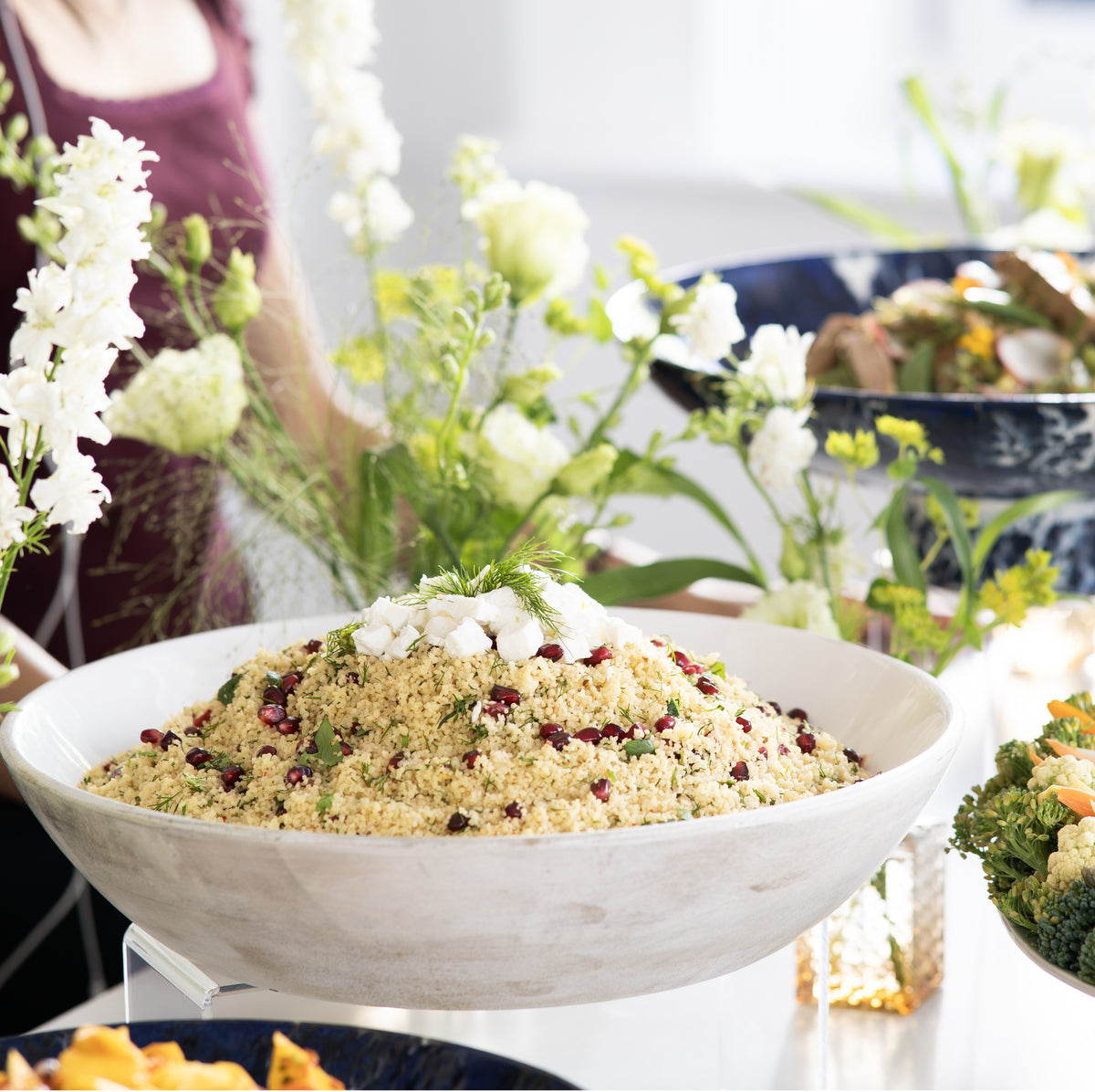 A large white oval serving bowl filled with a light beige couscous or grain salad, garnished with white cheese and herbs.
