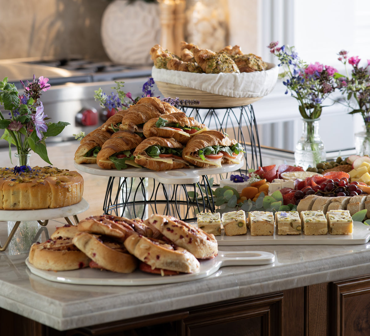 A buffet-style food spread on a marble counter, featuring croissants and sandwiches on white platters and stands, sliced fruit, crackers, and small cakes, with flowers and kitchen decor in the background.