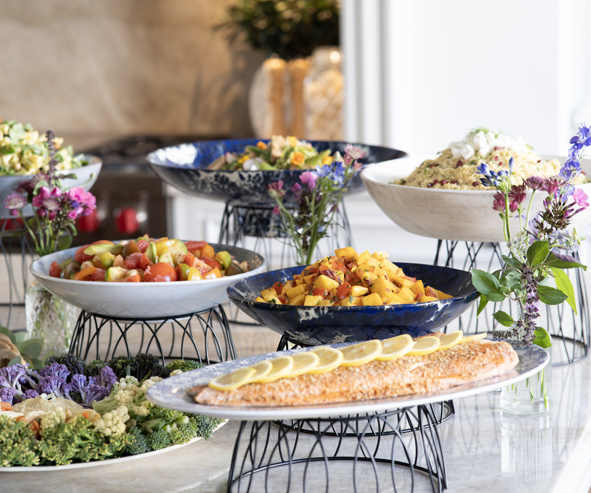 A lunch table with multiple bowls and platters of food on black wire stands, including a salmon fillet with lemon slices, mixed salads, and a grain bowl, set in a bright indoor environment.