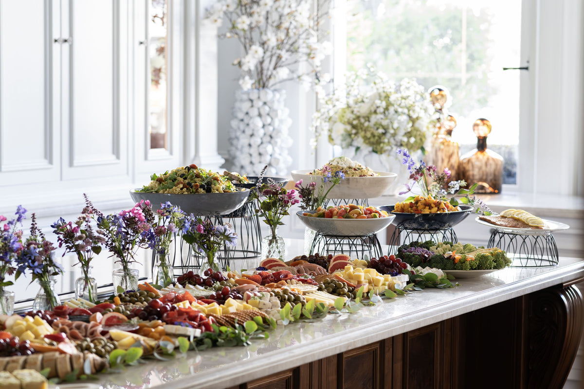 A long buffet table for office catering in london filled with assorted foods, salads, fruit, cheeses, and small floral arrangements.