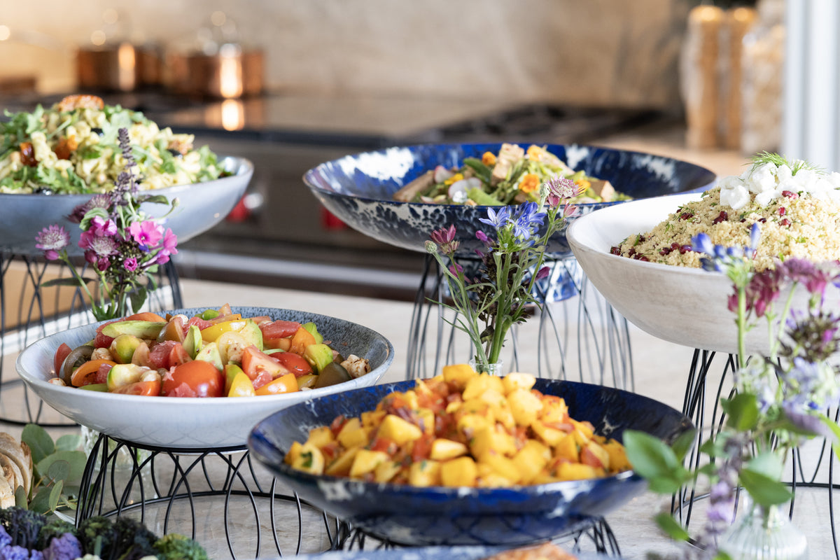 A buffet-style food display with multiple bowls of salads and fruit on wire stands in a kitchen or dining setting. 